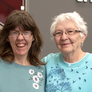 a woman in her 30s with brown hair and glasses stands next to her mom in her 70s with white hair and glasses