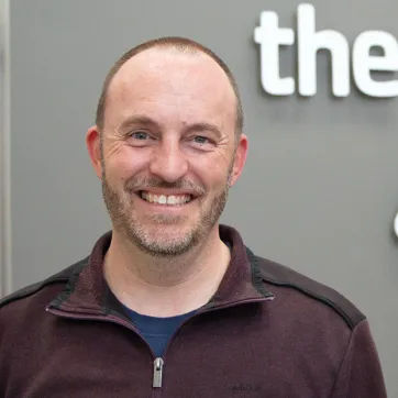 a photo of a man in a maroon zip jacket and light beard stubble smiles at the camera